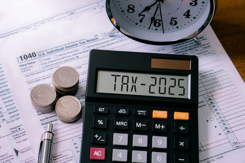 Calculator displaying 'TAX-2025' on a screen, surrounded by a stack of coins, a clock, and a 1040 U.S. Individual Income Tax Return form, symbolizing 2025 tax preparation