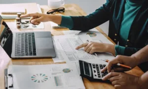 Two individuals working on financial tasks at a desk, with one person pointing at a laptop screen and the other using a calculator. Documents with charts, graphs, and data are spread across the table, accompanied by stacks of coins and an abacus in the background.