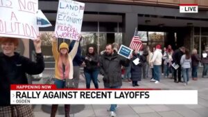 A group of protesters gathered outside a building, holding signs and American flags during a rally against recent layoffs in Ogden. The signs express concerns about job losses, economic impact, and federal worker rights. A news banner at the bottom of the image reads, "Happening Now: Rally Against Recent Layoffs - Ogden
