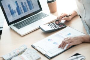 A financial professional working at a desk with a laptop displaying bar graphs, a calculator, a clipboard with financial charts, and stacks of cash.